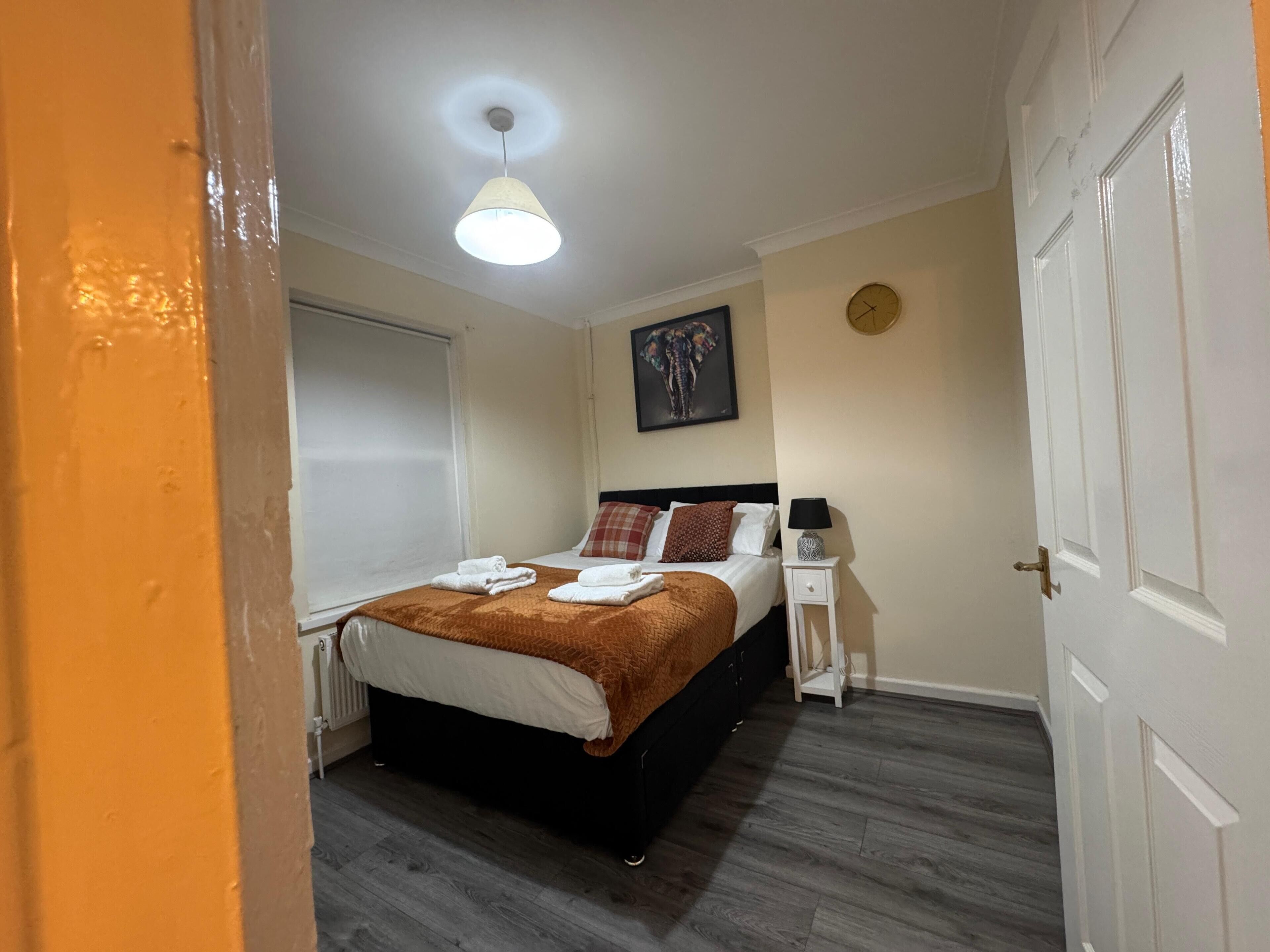 A bedroom with a bed, a bedside table, a wall clock, and a window with blinds.