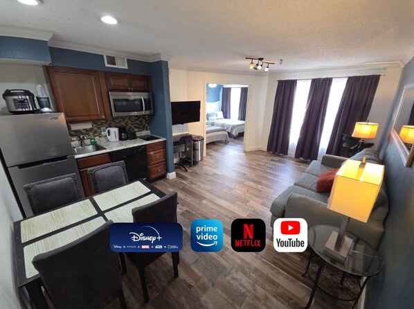 A modern hotel room with a kitchenette, a dining area, a living room with a sofa and a TV, and a bedroom with a bed and curtains.