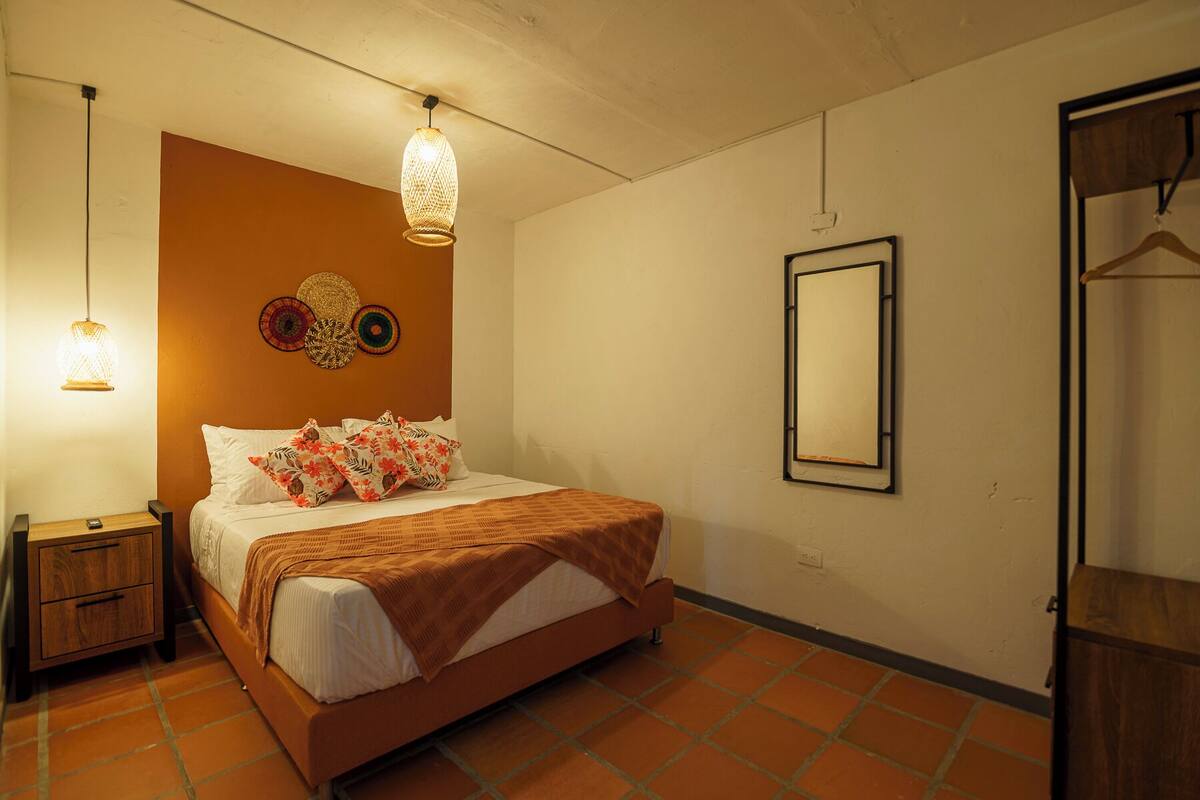 A bedroom with a bed, bedside tables, a hanging light fixture, a wall-mounted mirror, and a door.