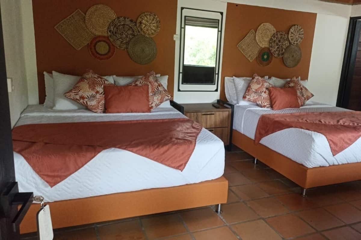 A hotel room with two beds, each with a brown blanket and white bedding. The room has a tiled floor and a window with a view of greenery outside.