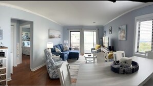 A spacious living room with a large window, a dining area, and a kitchen visible through an open door. - Beautiful 2-bedroom condo with ocean views, Wi-Fi, AC, wshr/dryr in Myrtle Beach (Myrtle Beach)