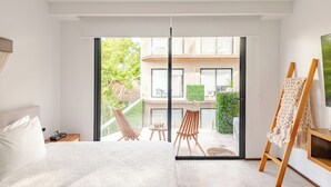 A modern hotel room with a large bed, a wooden nightstand, a desk with a chair, and a view of a balcony with outdoor furniture. - Tulum City Apartments M-209 (Tulum)
