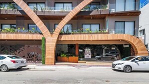 A modern building with a distinctive curved wooden facade, multiple balconies with plants, and a visible entrance with a sign. - Tulum City Apartments M-209 (Tulum)