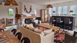 A cozy living room with a sofa, chairs, a wooden table, and a fireplace. There is a kitchen area with bar stools and a large window.