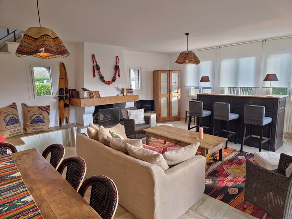 A cozy living room with a sofa, chairs, a wooden table, and a fireplace. There is a kitchen area with bar stools and a large window.