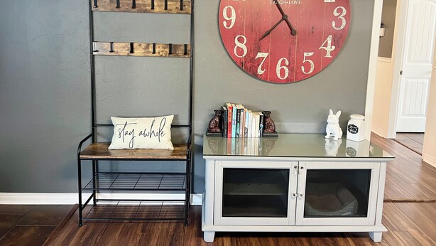 Ein Zimmer mit einer großen roten Uhr, einem Metallregal, einem weißen Schrank mit Glastüren und einem Kissen mit dem Schriftzug 'stay awhile'.
