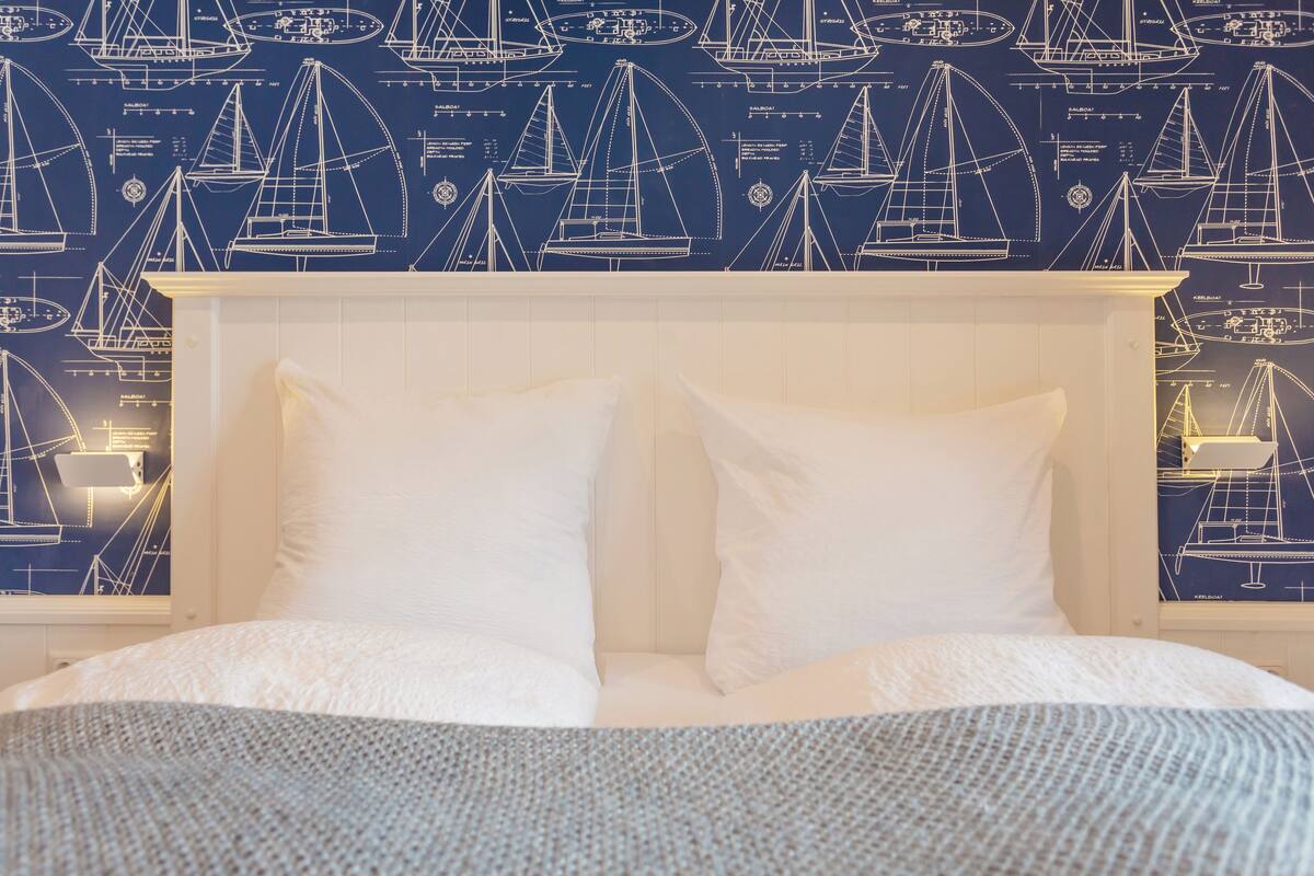 Un lit avec une literie blanche, dans une chambre dont le papier peint est décoré sur le thème de la marine, avec des motifs de voiliers et des schémas de navires.