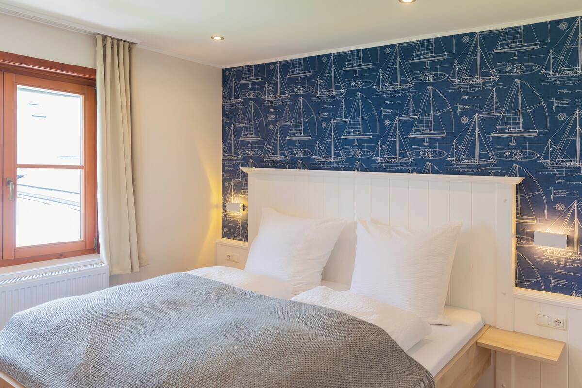 Une chambre à coucher comprenant un lit, une table de chevet, une fenêtre avec des rideaux et un mur recouvert d’un papier peint sur le thème de la marine.