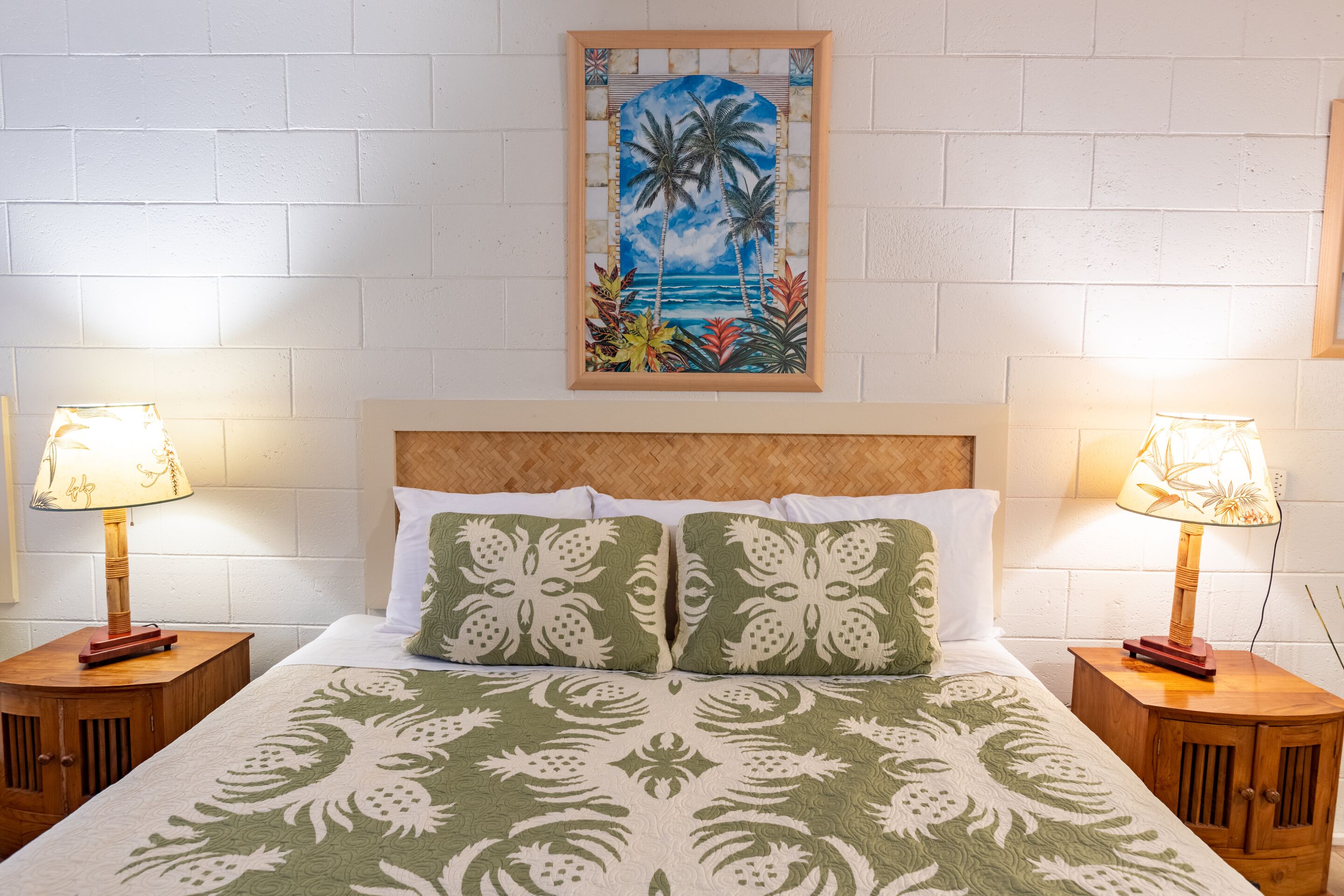 Un lit bien fait, recouvert d’une couvre-lit à motifs floraux verts, avec deux oreillers assortis, et un tableau représentant un paysage tropical encadré au-dessus. De chaque côté du lit, on trouve deux tables de chevet en bois sur lesquelles sont posées des lampes.