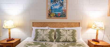 A neatly made bed with a green floral bedspread, two matching pillows, and a framed tropical painting above. There are two wooden nightstands with lamps on either side of the bed.