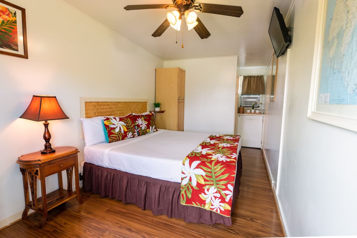 Habitación de hotel con cama, mesita de noche de madera con lámpara, ventilador de techo, área de cocina al fondo y televisor montado en la pared.