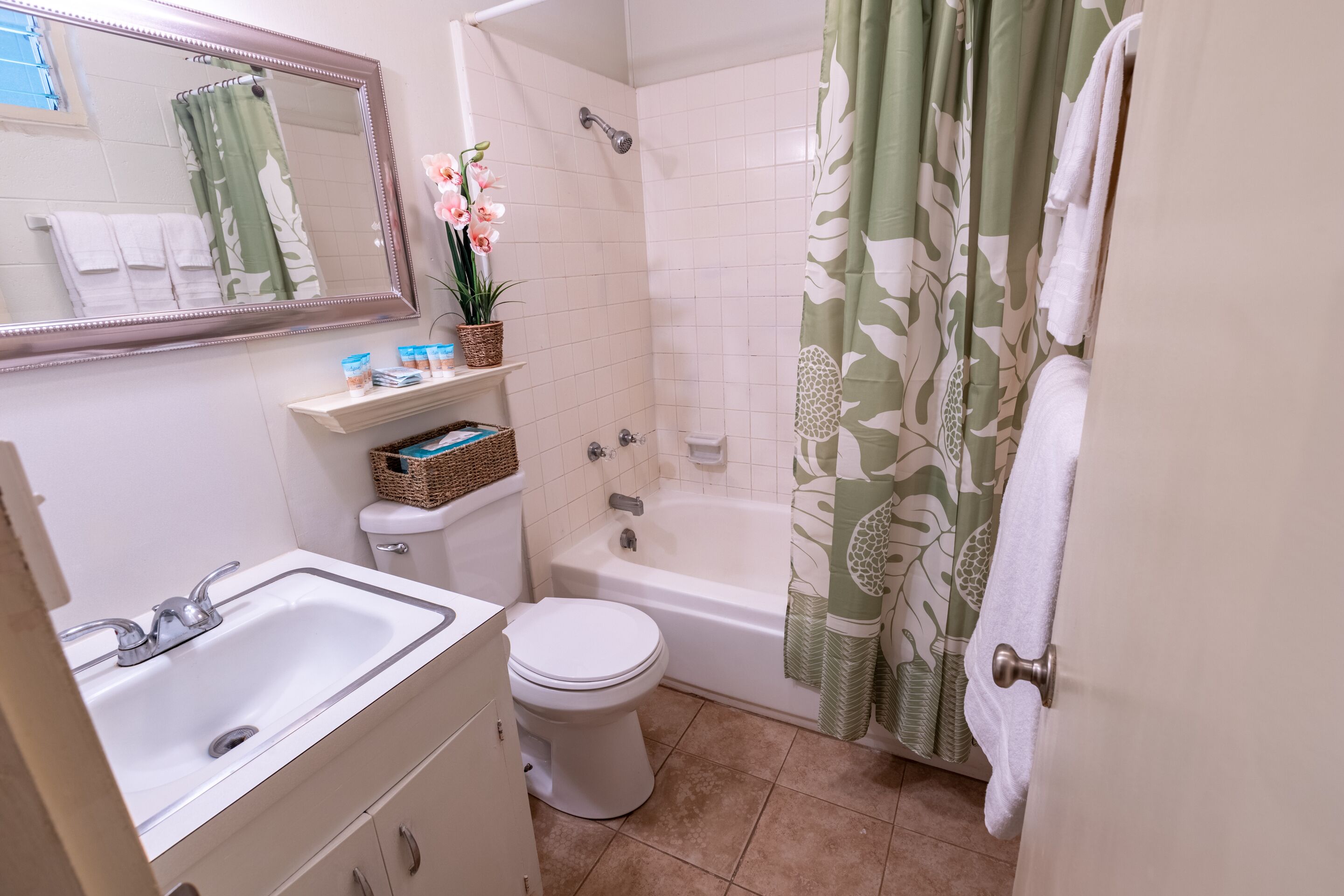Baño con lavamanos, inodoro y bañera. Hay un espejo sobre el lavamanos y una cortina de ducha con estampado floral.
