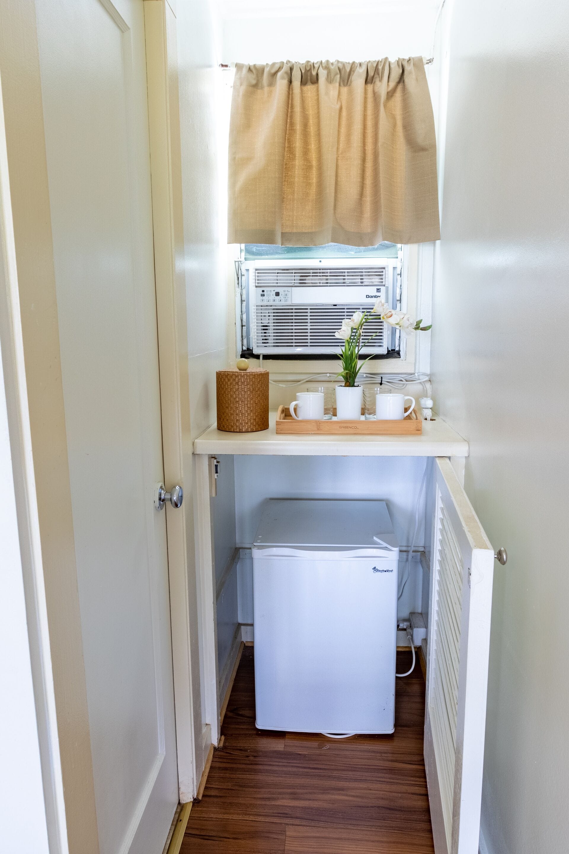 Una kitchenette compacta con un refrigerador pequeño, una ventana con cortina y una planta en maceta sobre la encimera.