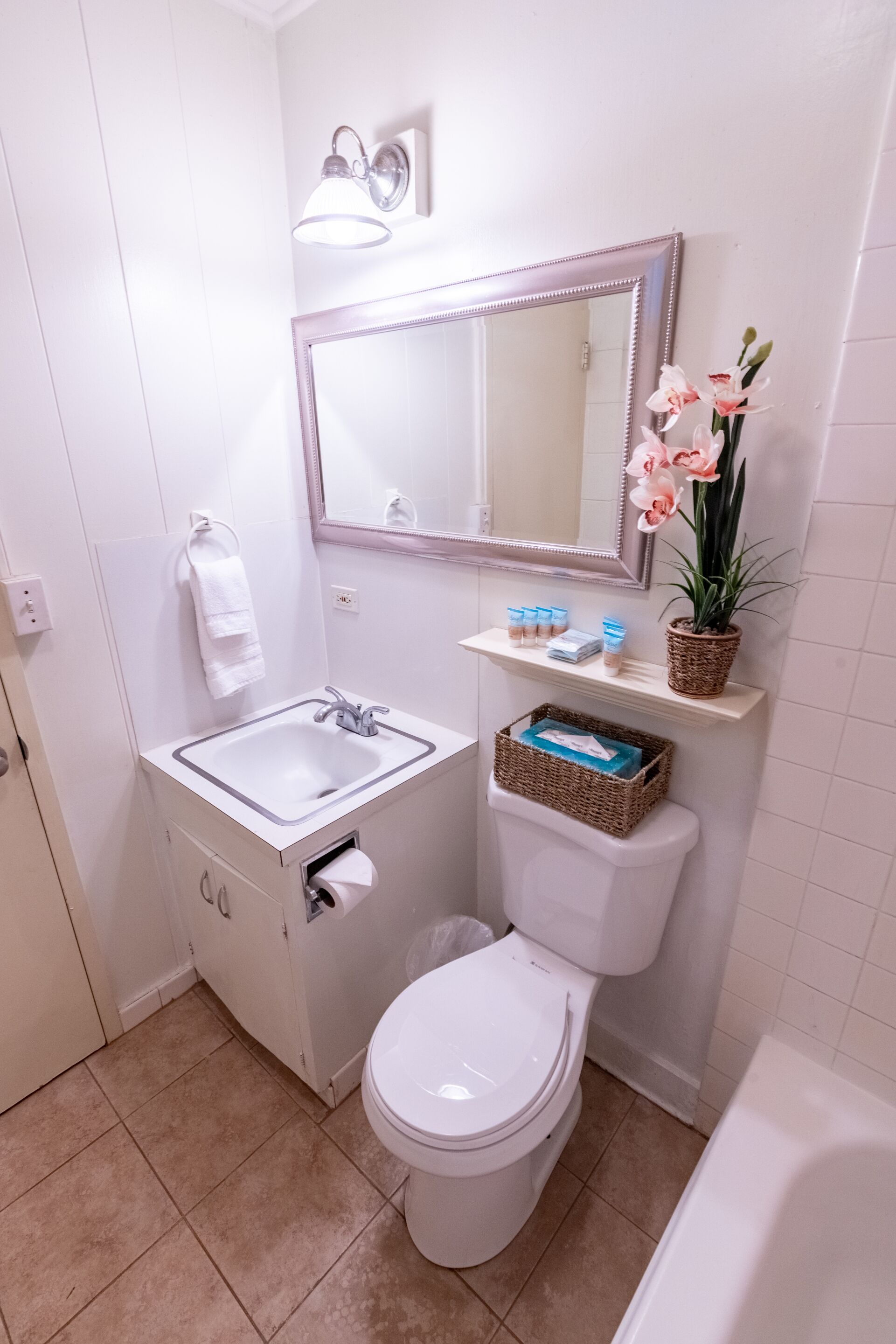 Un baño con lavamanos, inodoro y bañera blancos. Hay un espejo sobre el lavamanos y una repisa con artículos de aseo.
