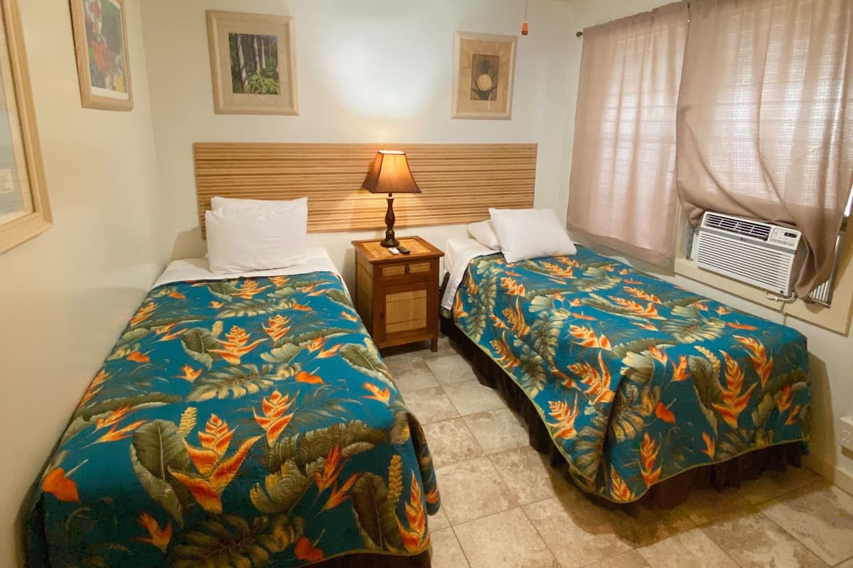 Habitación de hotel con dos camas, cada una con cubrecama floral, cabecero de madera, mesita de noche con lámpara y ventana con cortinas.