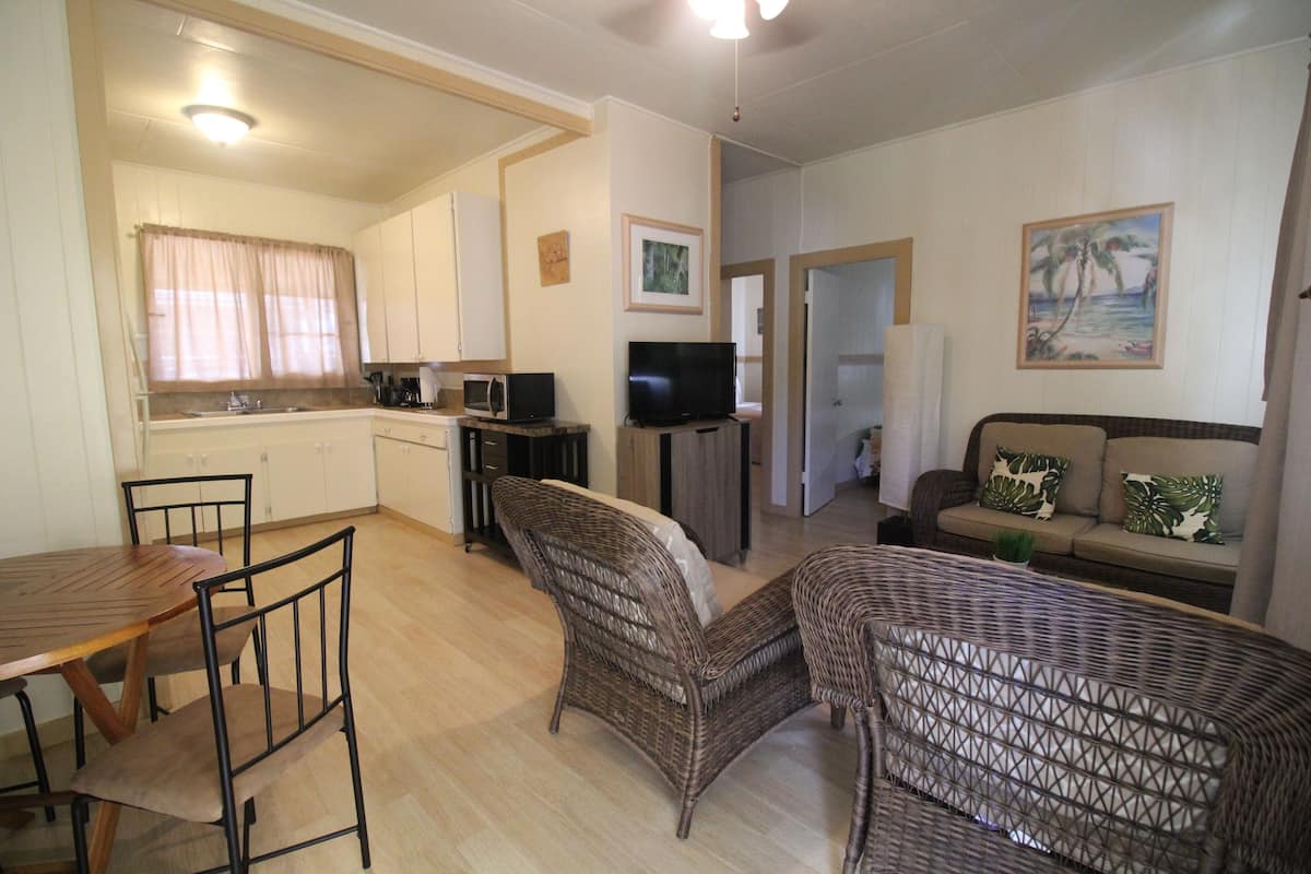 Un espacio habitable compacto con comedor, kitchenette y zona de estar con sofá y dos sillones de mimbre.