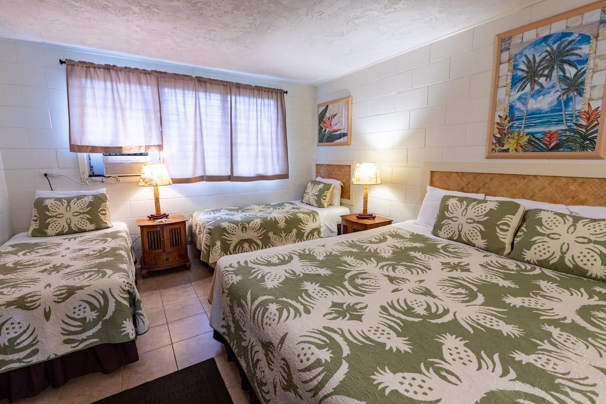 Habitación de hotel con dos camas individuales y una cama doble, cada una con ropa de cama de estampado floral a juego. Hay dos mesitas de noche con lámparas, una ventana con cortinas y un cuadro con temática tropical en la pared.