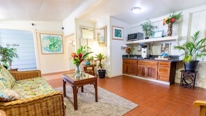 A cozy living room with a wicker sofa, a glass-top coffee table, and a kitchen area with wooden cabinets and a microwave. - Kaua'i Palms Hotel (Lihue)