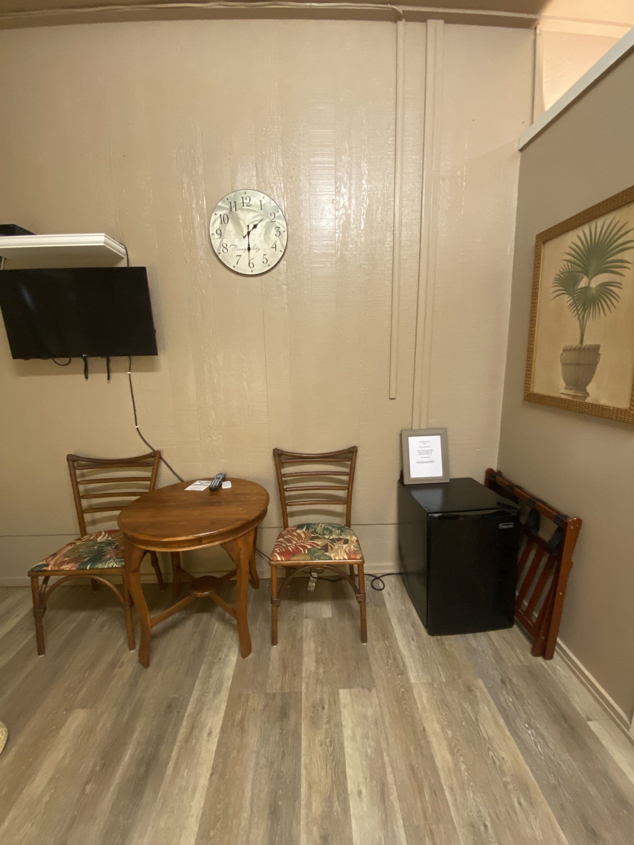 A room with a wooden dining table, two chairs, a wall-mounted clock, a flat-screen TV, a small black refrigerator, and a framed plant on the wall.