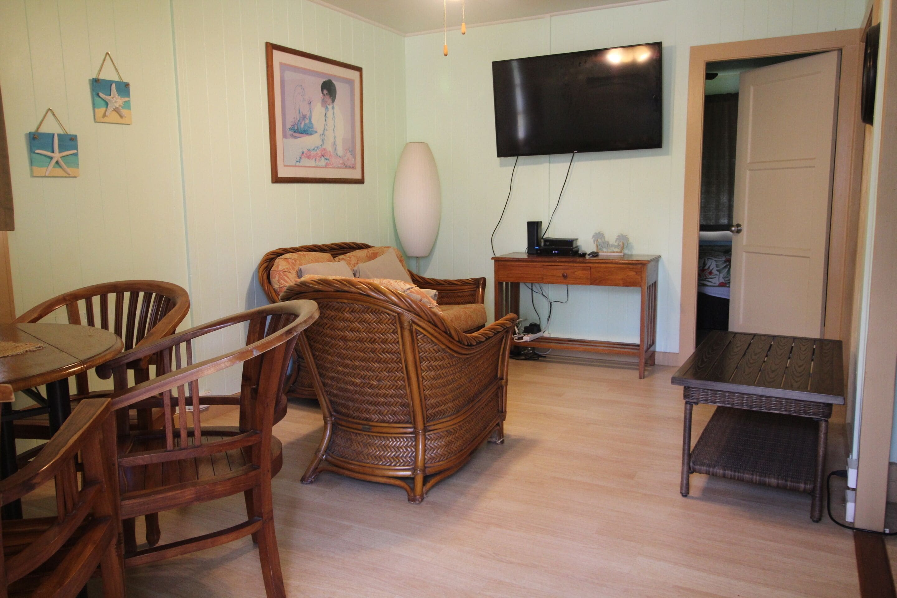 A living room with a wicker sofa, wooden chairs, a small table, a TV mounted on the wall, and a door leading to another room.