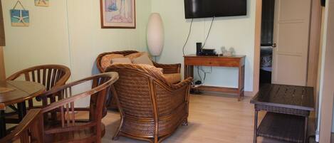A living room with a wicker sofa, wooden chairs, a small table, a TV mounted on the wall, and a door leading to another room.