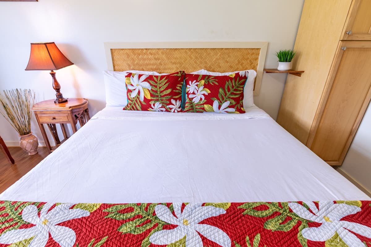 Una cama bien tendida con edredón blanco y colcha floral roja. Se observa un cabecero de madera, una mesita de noche con lámpara y una pequeña estantería con una planta.