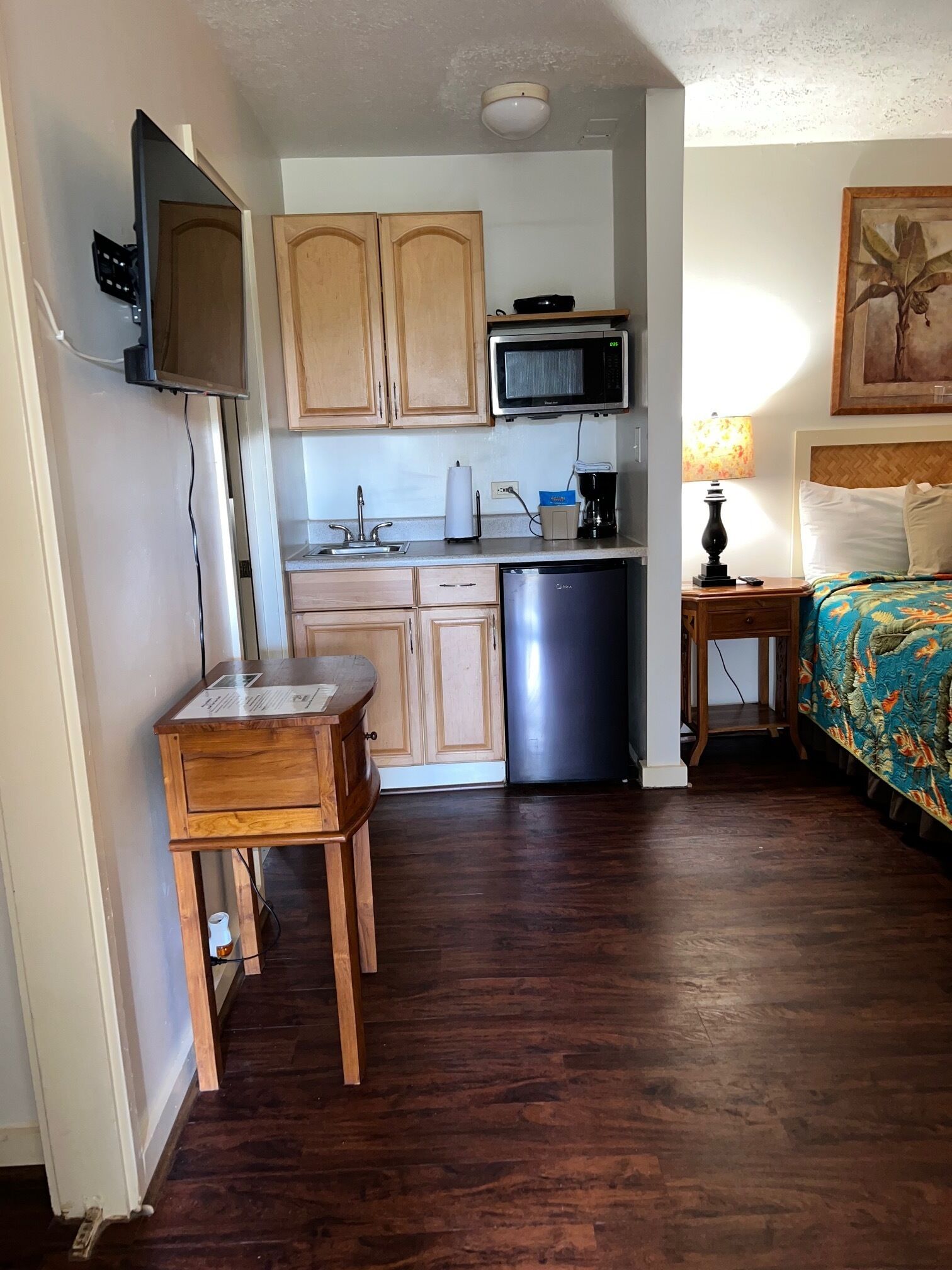 Une cuisine compacte avec des meubles en bois, un four à micro-ondes et un lave-vaisselle. La pièce adjacente contient une petite table en bois et un lit recouvert d’une courtepointe aux couleurs vives.