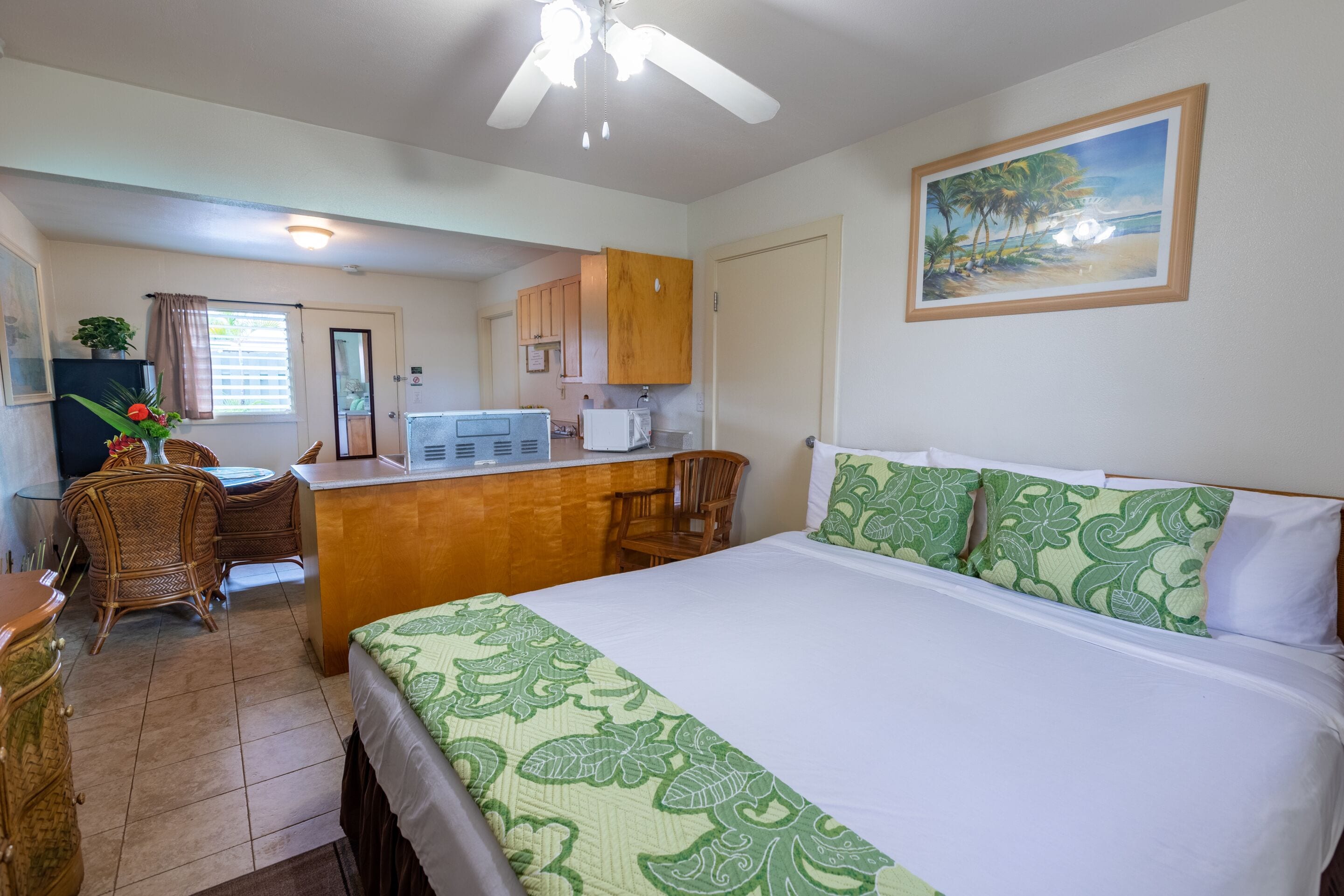 A hotel room with a bed, a desk, a chair, a kitchenette, and a painting on the wall.