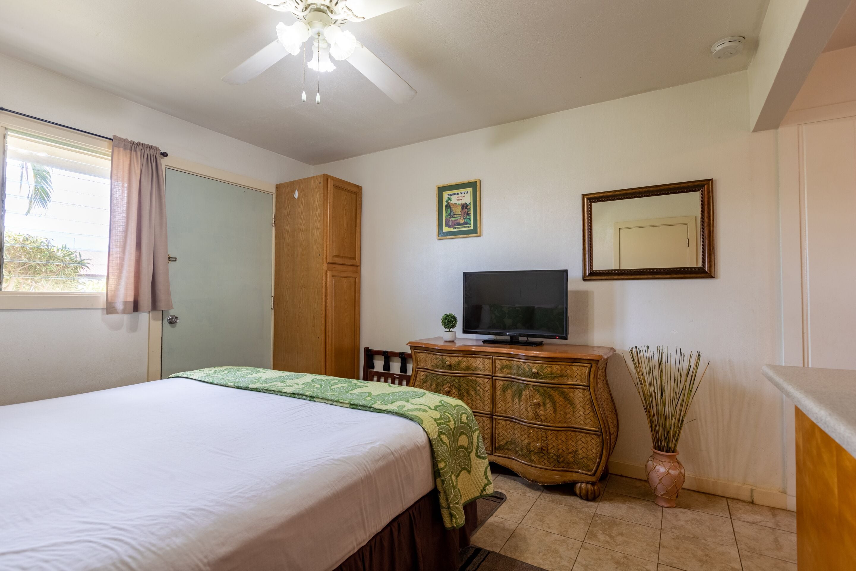 A bedroom with a bed, a television on a wooden dresser, a ceiling fan, and a window with curtains.