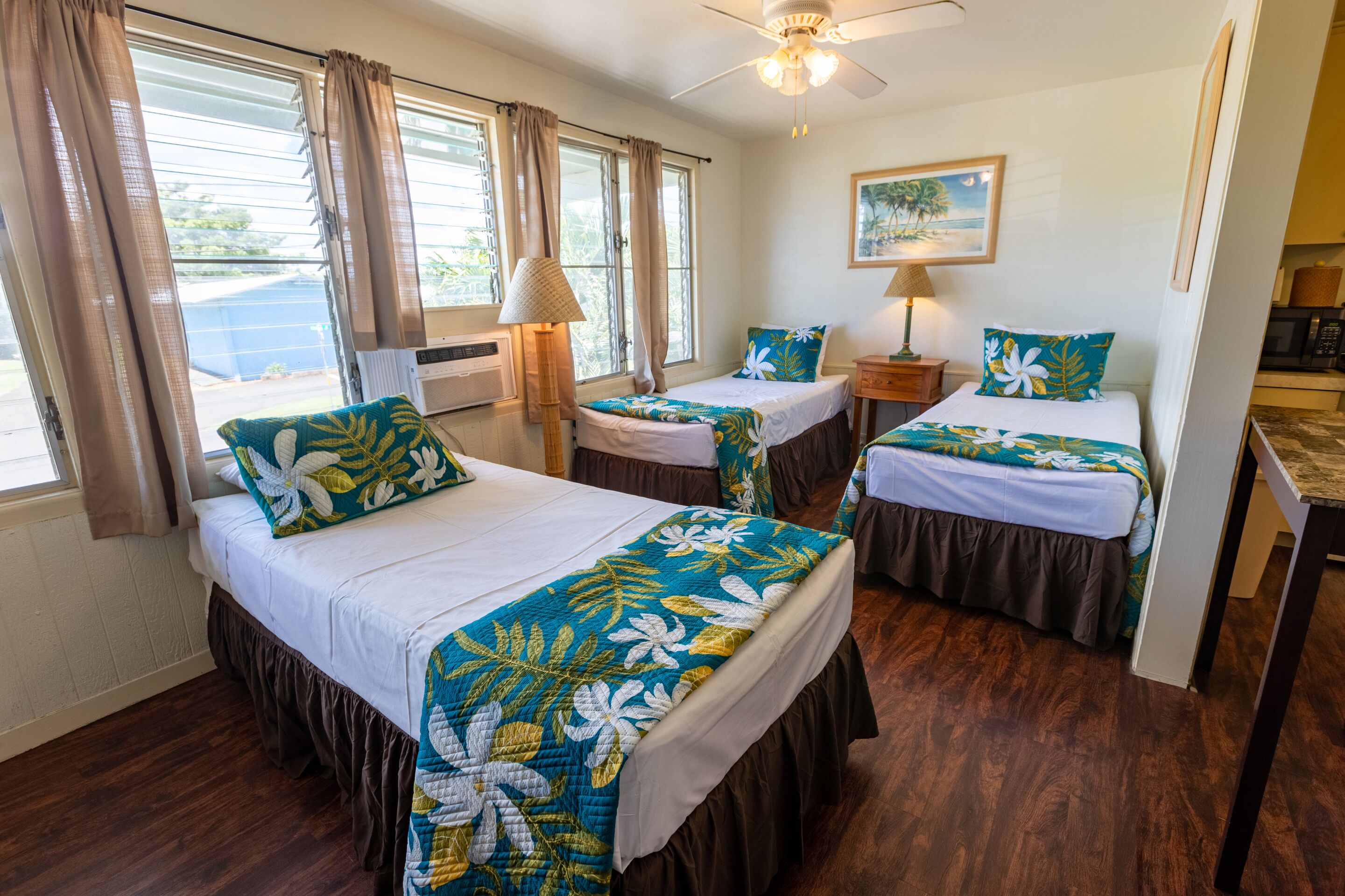 A hotel room with two single beds, a ceiling fan, a painting on the wall, and a view of the outdoors through the windows.