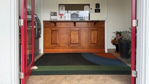 Zona de recepción de hotel con mostrador de madera, una puerta con marco rojo, felpudo de bienvenida y un tablón de anuncios con varios carteles.