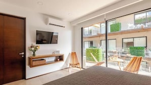 A modern bedroom with a large bed, a TV mounted on the wall, and a balcony with outdoor seating. - Tulum City Apartments M-221 (Tulum)