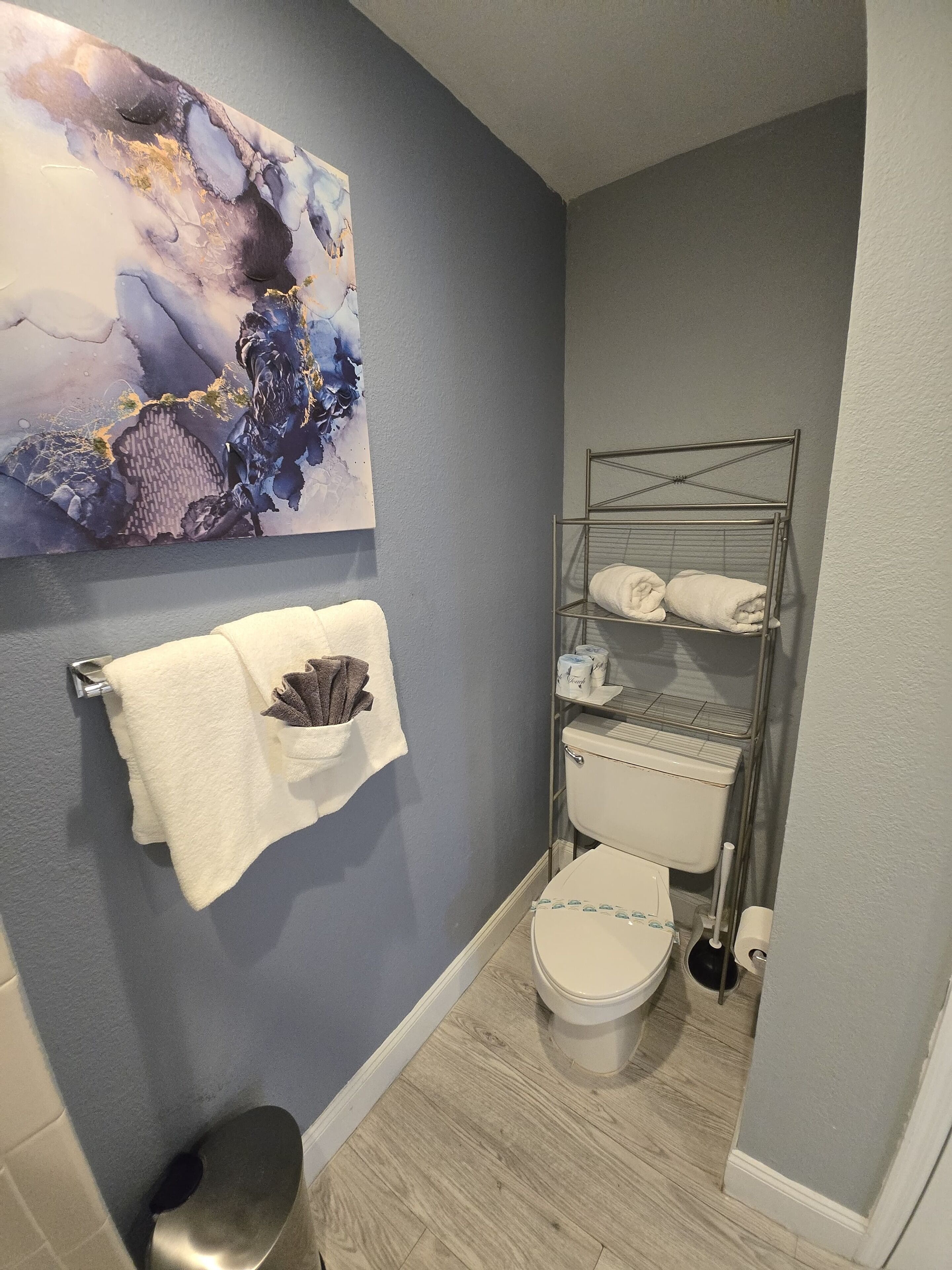 A bathroom with a toilet, a metal towel rack, a wall-mounted towel dispenser, and a large abstract painting.