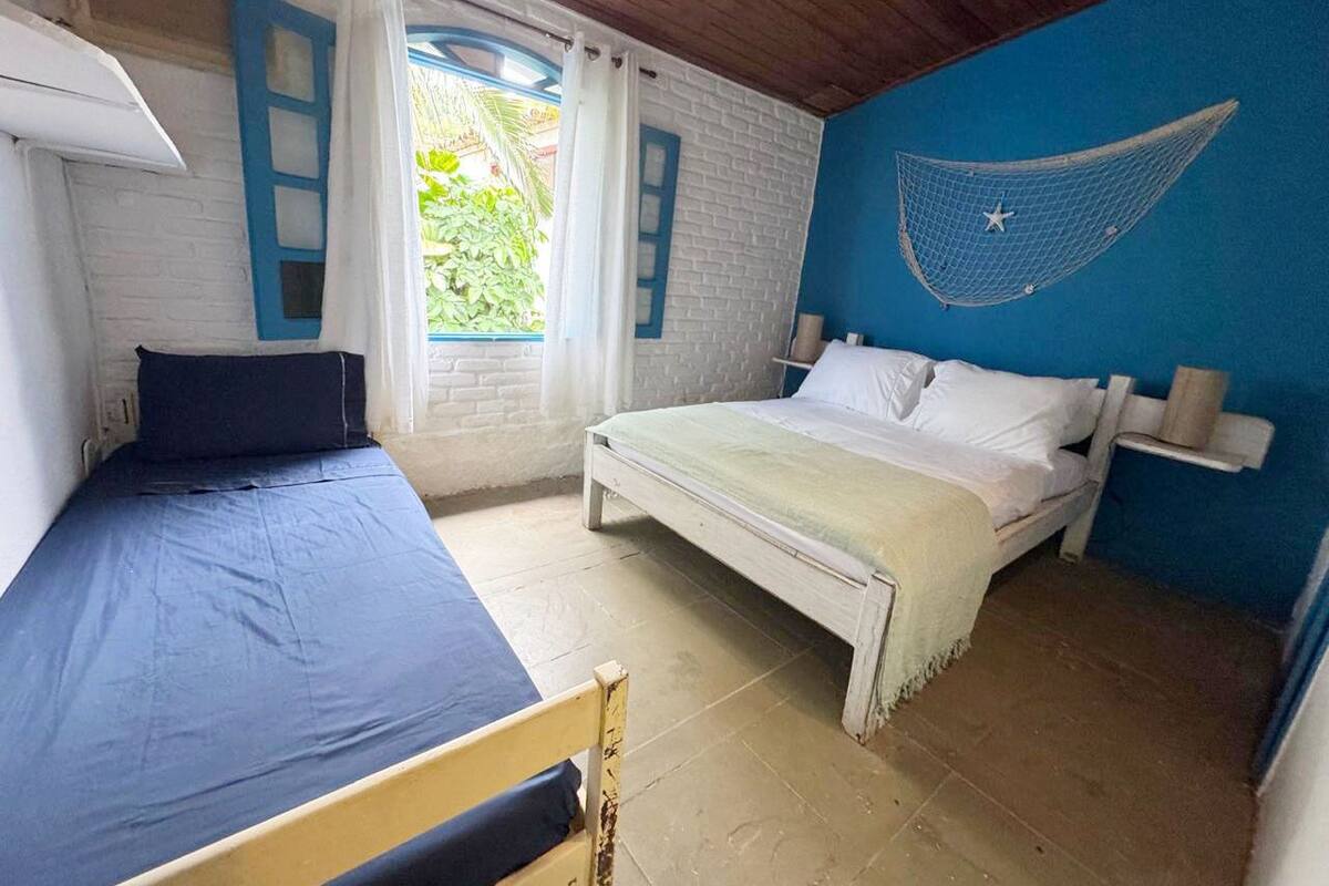Ein Zimmer mit zwei Betten, eines mit blauer Bettdecke und das andere mit weißer Bettdecke. Eine Hängematte ist an der Wand befestigt und es gibt ein Fenster mit Vorhängen.