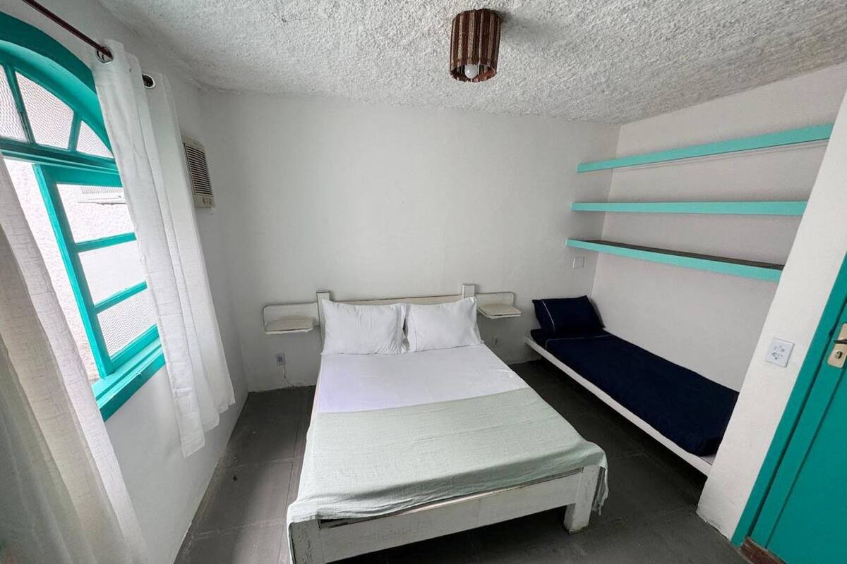 Ein kleines, schlichtes Zimmer mit einem Bett, einem Vorhangfenster, einem Regal und einer Tür.