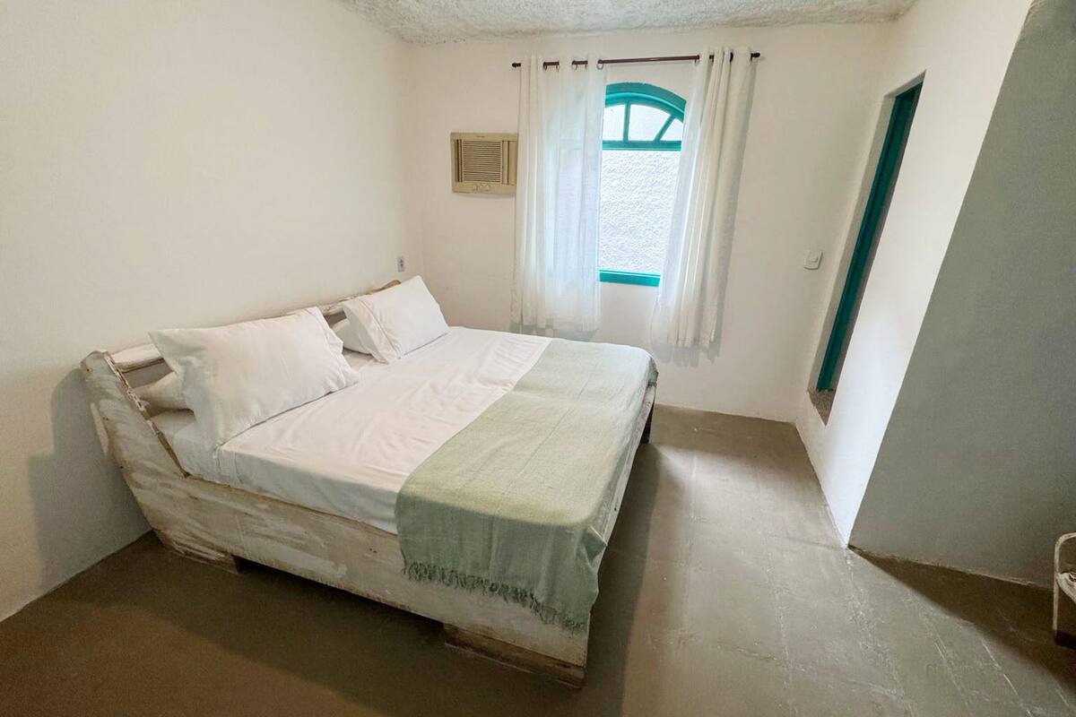 Ein Zimmer mit einem Einzelbett, schlicht eingerichtet, mit einem Fenster mit Vorhängen und einer kleinen Wand-Klimaanlage.