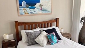 A neatly made bed with a wooden headboard, a painting of a coastal scene above it, and a small bedside table with a lamp. - Ocean views from your spacious balcony at Seaside Escape Aria (Rockingham)
