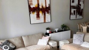 A modern living room with a beige sofa, a round ottoman, and two side tables. There are abstract paintings on the wall and a potted plant on the side table. - Ocean views from your spacious balcony at Seaside Escape Aria (Rockingham)
