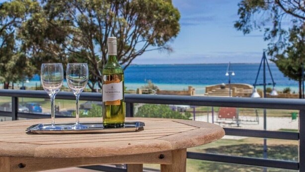 A round wooden table with a bottle of wine and two glasses on a tray, overlooking a beach with trees and the ocean. - Ocean views from your spacious balcony at Seaside Escape Aria (Rockingham)