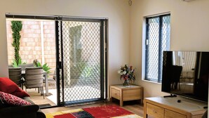 A living room with a sofa, a TV, a wooden cabinet, a colorful rug, a small table with a vase, and a sliding glass door leading to an outdoor area with a brick wall and plants. - Victoria Views - Your home away from home (Rockingham)
