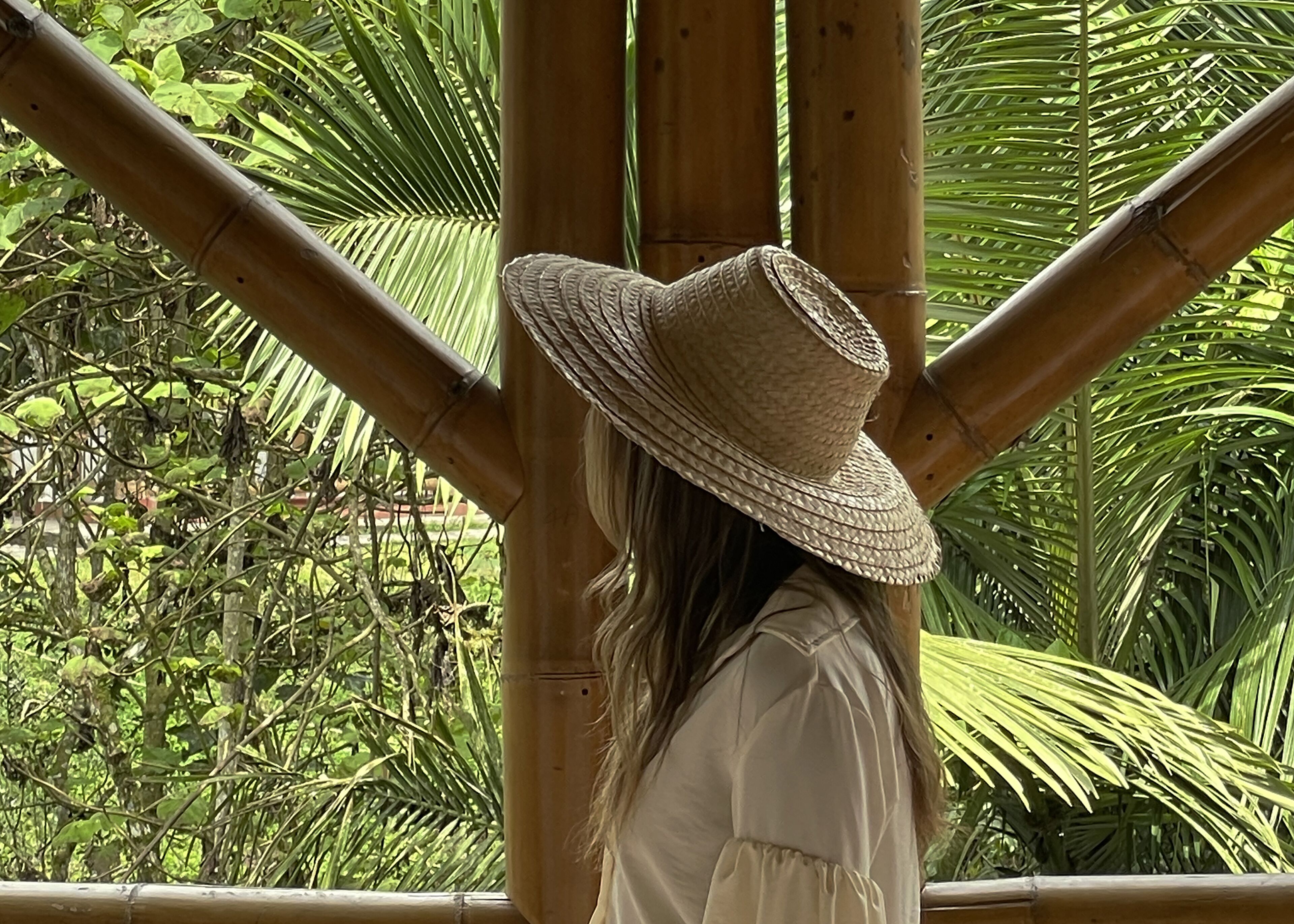 Uma pessoa usando chapéu de palha em frente a uma estrutura de bambu, com folhagem verde exuberante ao fundo.