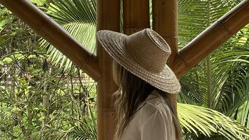 Uma pessoa usando chapéu de palha em frente a uma estrutura de bambu, com folhagem verde exuberante ao fundo.