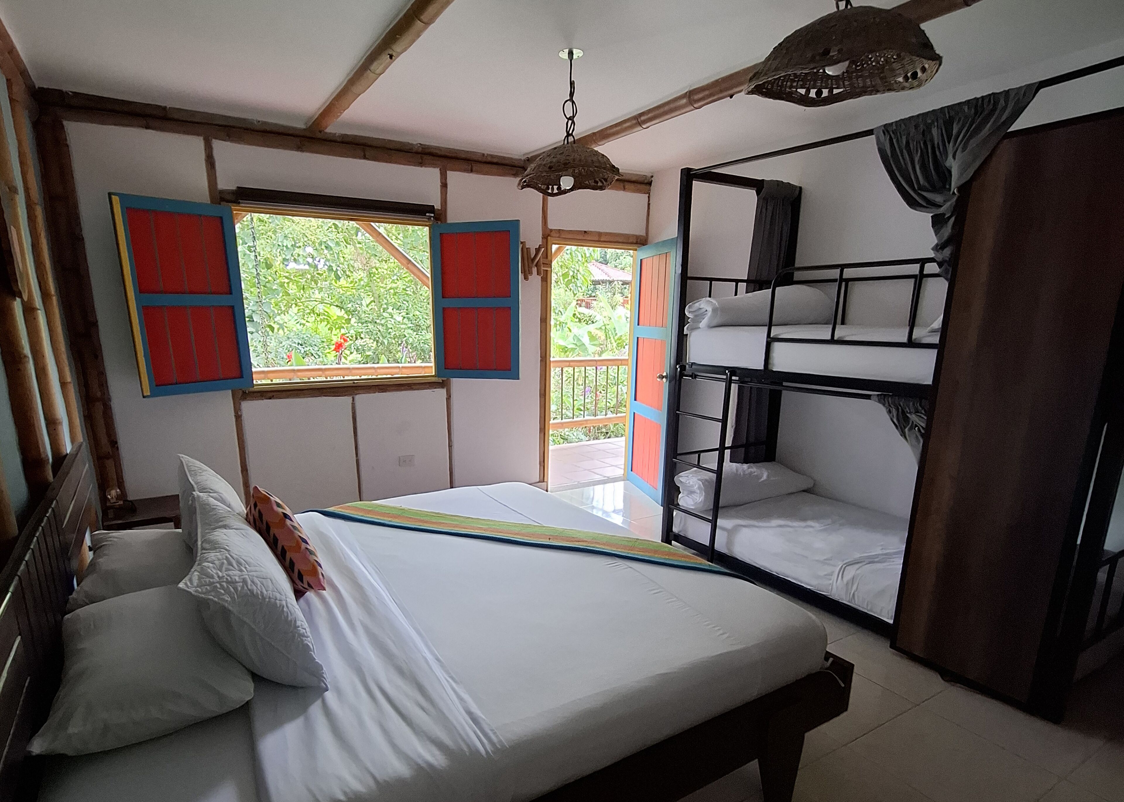 Une chambre avec un lit simple, un lit superposé, une fenêtre avec des volets rouges et un luminaire suspendu.