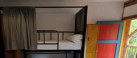 Una cama litera con cortina, un armario de madera con paneles rojos y azules, y una lámpara de fibra trenzada.