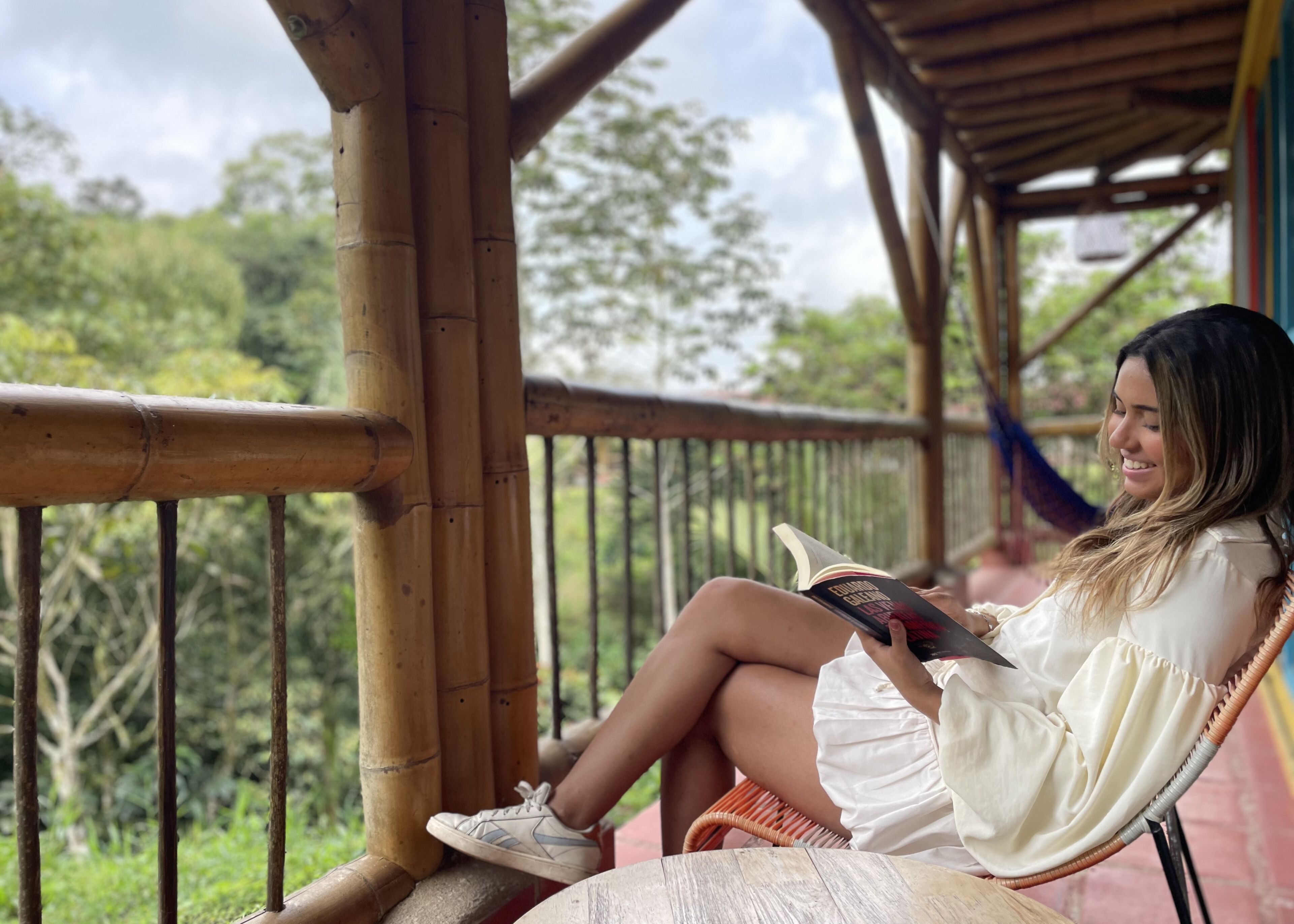 Una persona sentada en una silla de mimbre leyendo un libro en una terraza de madera con barandas de bambú, rodeada de abundante follaje.