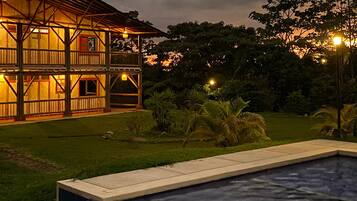 Una casa de madera de dos pisos con un balcón, rodeada por un césped cuidado y una piscina.