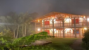 Un edificio de dos pisos con una estructura de balcón de madera, rodeado de abundante vegetación.