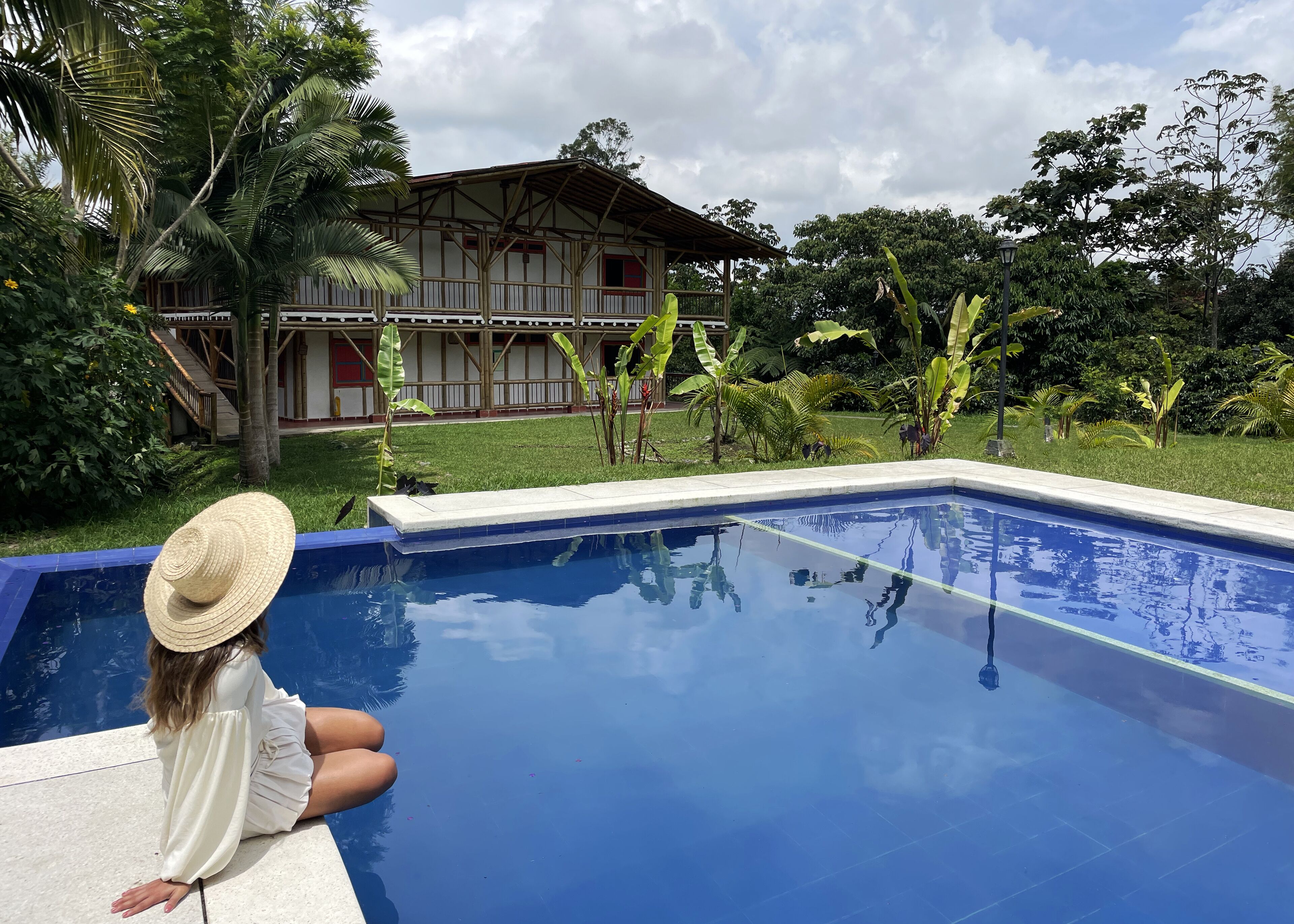Una piscina con una persona sentada junto a ella, rodeada de exuberante vegetación y un edificio de madera.