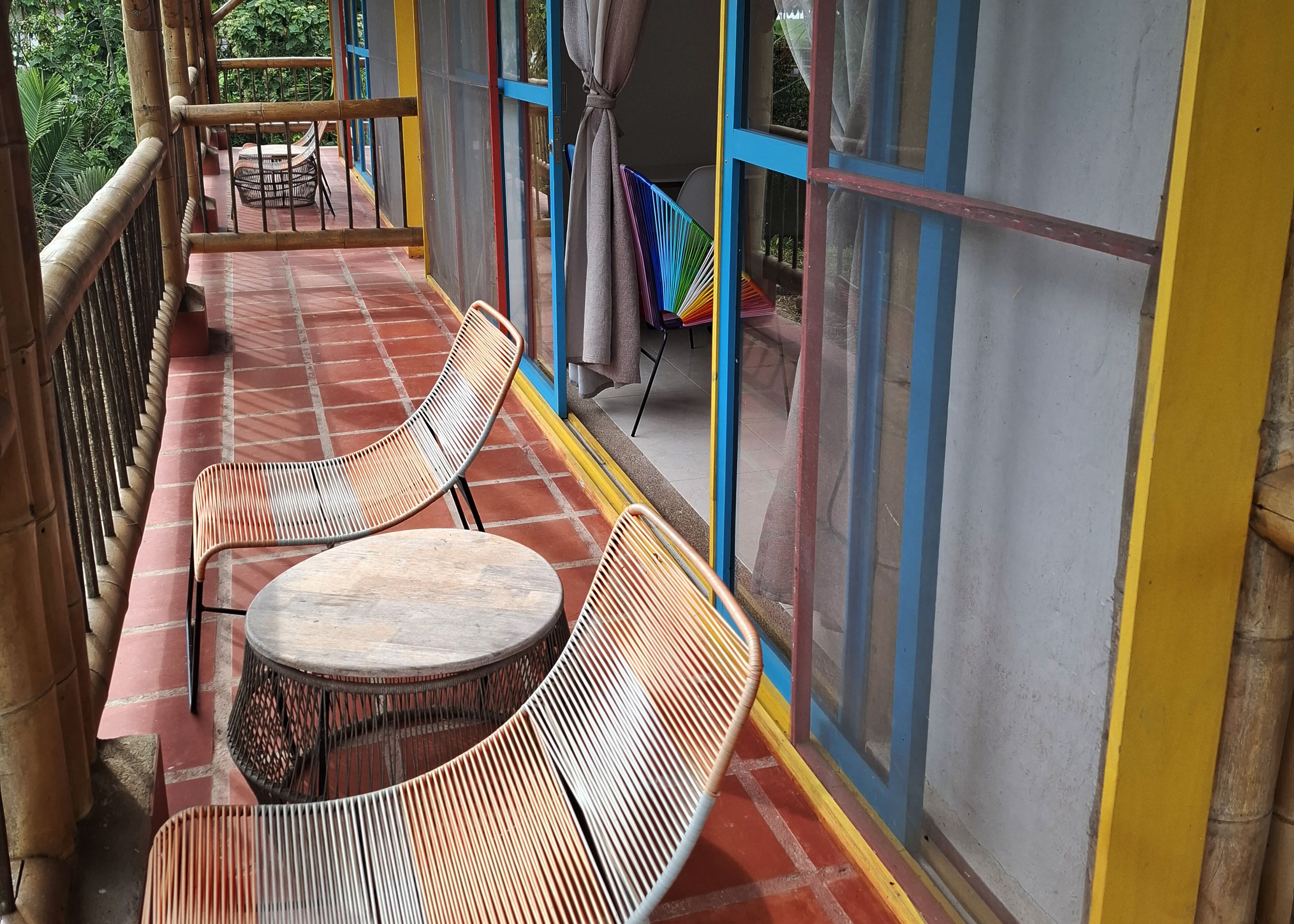 A covered outdoor seating area with two round tables and wicker chairs, a view of greenery through large windows, and a bamboo railing.