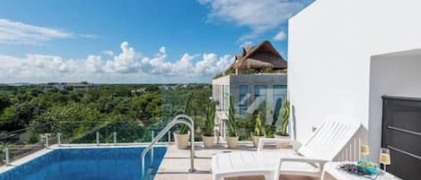 A modern pool area with a clear view of the surrounding landscape, a thatched-roof structure, and a white building with a balcony.